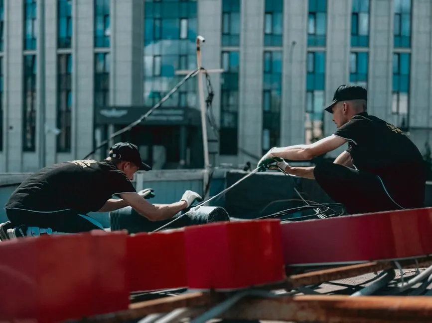 Two workers are engaged in maintenance work on a rooftop against a city building backdrop.